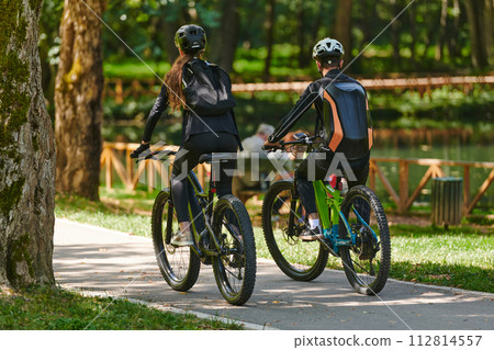 A blissful couple, adorned in professional cycling gear, enjoys a romantic bicycle ride through a park, surrounded by modern natural attractions, radiating love and happiness A blissful couple, adorned in professional cycling gear, enjoys a romantic bicycle ride through a park, surrounded by modern natural attractions, radiating love and happiness 112814557