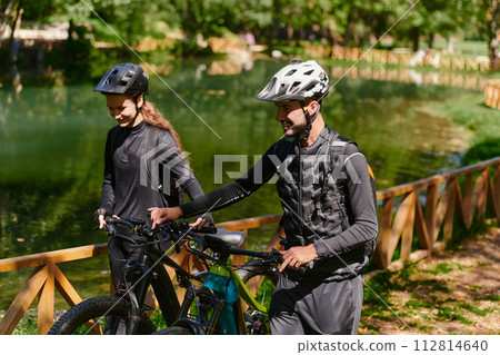 A blissful couple, adorned in professional cycling gear, enjoys a romantic bicycle ride through a park, surrounded by modern natural attractions, radiating love and happiness 112814640