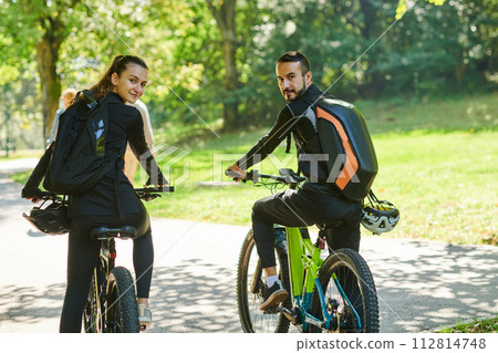 A blissful couple, adorned in professional cycling gear, enjoys a romantic bicycle ride through a park, surrounded by modern natural attractions, radiating love and happiness 112814748