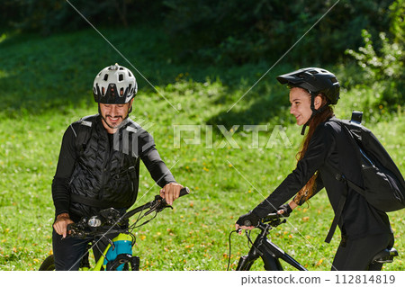 A blissful couple, adorned in professional cycling gear, enjoys a romantic bicycle ride through a park, surrounded by modern natural attractions, radiating love and happiness 112814819