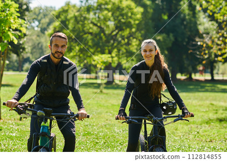A blissful couple, adorned in professional cycling gear, enjoys a romantic bicycle ride through a park, surrounded by modern natural attractions, radiating love and happiness 112814855