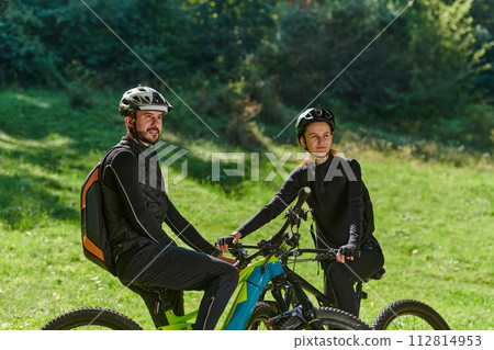 A sweet couple, equipped with bicycles and engrossed in coordinating their journey, checks their GPS mobile and watches while planning scenic routes in the park, seamlessly blending technology and 112814953