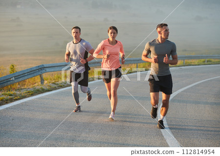 A group of friends, athletes, and joggers embrace the early morning hours as they run through the misty dawn, energized by the rising sun and surrounded by the tranquil beauty of nature 112814954