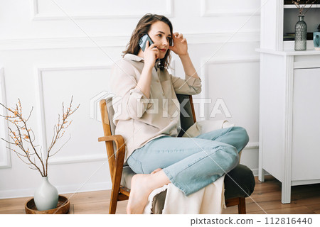 Serious millennial woman talks on the phone, sitting on a chair and looking out the window in her 112816440