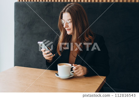 Young happy businesswoman smiling and using smart phone in a cafe. Person sitting at table and using smartphone indoors. Online education, order, working or shopping concept 112816543