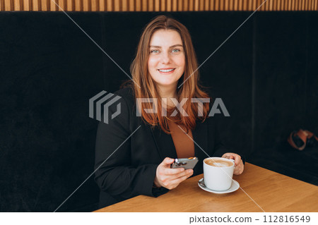 Young happy businesswoman smiling and using smart phone in a cafe. Person sitting at table and using smartphone indoors. Online education, order, working or shopping concept Young happy businesswoman smiling and using smart phone in a cafe. Person sitting at table and using smartphone indoors. Online education, order, working or shopping concept 112816549