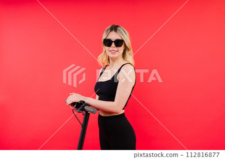 Blonde woman on red white background sitting on electro scooter and resting, copy space Blonde woman on red white background sitting on electro scooter and resting, copy space 112816877