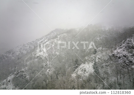 Aerial foggy landscape with mountain cliffs covered with fresh fallen snow during heavy snowfall in winter mountain forest on cold quiet day 112818360