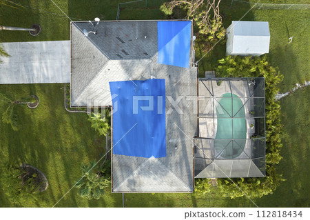 Aerial view of damaged in hurricane Ian house roof covered with blue protective tarp against rain water leaking until replacement of asphalt shingles 112818434