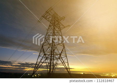 Transmission and long distance distribution of electricity concept. High voltage tower with electric power lines stretching on dark blue starry sky.and bright distant city lights background. Transmission and long distance distribution of electricity concept. High voltage tower with electric power lines stretching on dark blue starry sky.and bright distant city lights background. 112818467