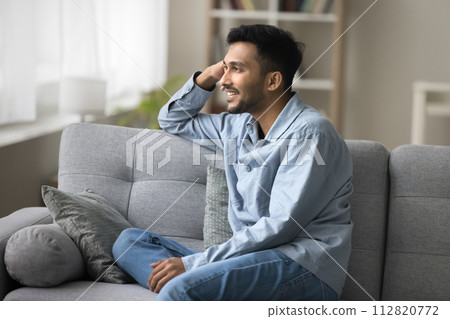 Indian guy relaxing seated on sofa smiling looking out window Indian guy relaxing seated on sofa smiling looking out window 112820772