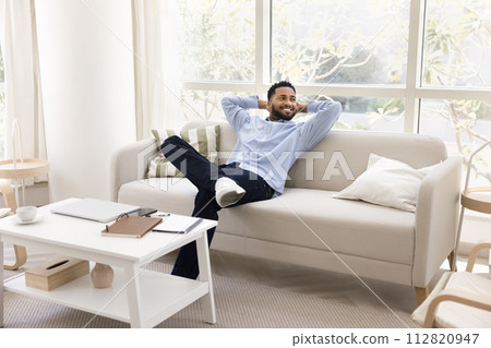Cheerful young black freelance employee man stretching body on couch Cheerful young black freelance employee man stretching body on couch 112820947