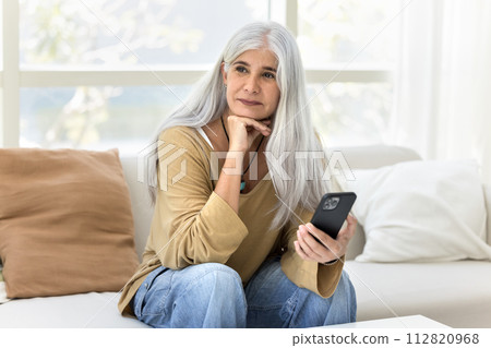 Thoughtful happy elder Latin woman holding smartphone 112820968