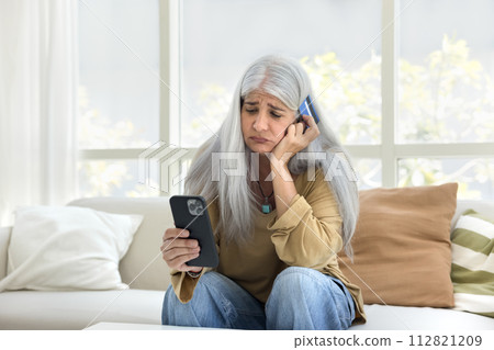 Sad elderly Hispanic woman looking at phone and credit card 112821209