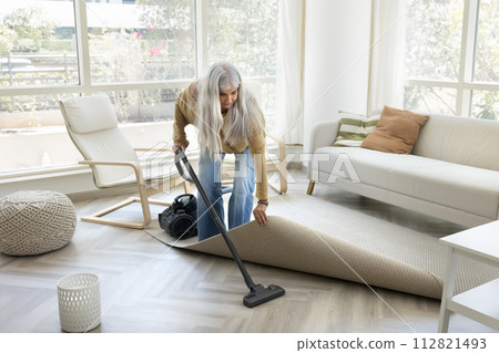 Serious grey haired mature housemaid woman hoovering floor under rug Serious grey haired mature housemaid woman hoovering floor under rug 112821493