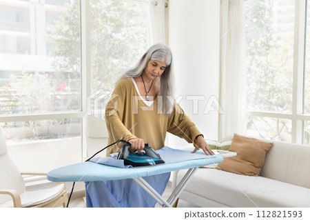 Serious senior Latin maid woman ironing clothing in living room Serious senior Latin maid woman ironing clothing in living room 112821593