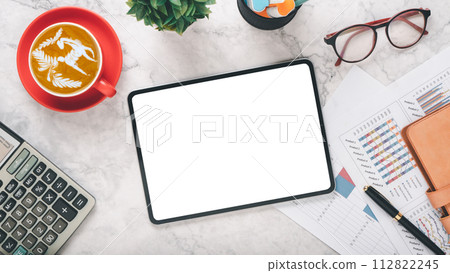 A professional workspace scene featuring a blank tablet screen, a cup of latte art coffee in a red mug, glasses, and financial charts, all arranged on a marble surface. 112822245