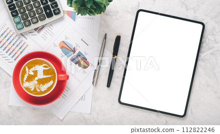A well organized desk featuring a tablet with a blank screen, artistic latte in a red cup, calculator, pen, and financial summaries on paper, all atop a polished marble background. 112822246