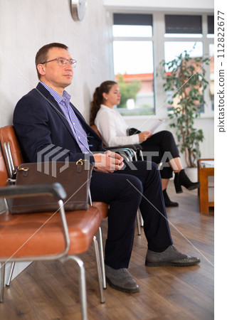 Serious-minded male candidate siting on chair and waiting for job interview in hallway Serious-minded male candidate siting on chair and waiting for job interview in hallway 112822677