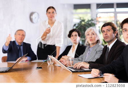 Office coworkers of different ages watching presentation while sitting at table with laptop and notes 112823038