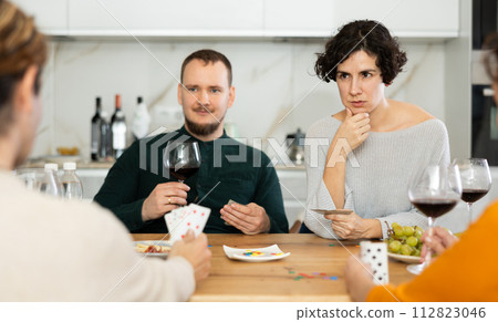 Friendly company of men and women sitting at kitchen table and enjoying the poker game. Friends play a card game at home. Evening entertainment concept Friendly company of men and women sitting at kitchen table and enjoying the poker game. Friends play a card game at home. Evening entertainment concept 112823046