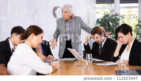 Outraged old female manager getting mad at office workers sitting with reports and laptops at table in meeting room Outraged old female manager getting mad at office workers sitting with reports and laptops at table in meeting room 112823047