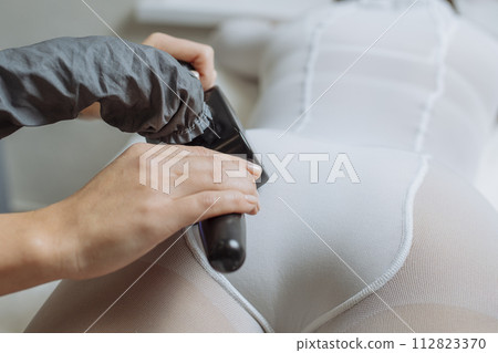 Close up shot of therapist beautician makes LPG massage treatment to young woman in white transparent suit, lying at beauty SPA clinic. Focus on hands holding medical unit 112823370