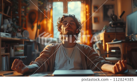 A man sits contentedly with eyes closed, soaking up the warm sunlight in his cozy, cluttered home office, exuding a sense of peace and relaxation. 112824385