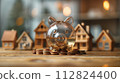 A transparent piggy bank filled with coins stands in focus, with blurred miniature houses in the background, symbolizing home loan real estate investment savings. 112824400
