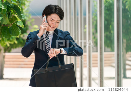 一位身穿西裝的年輕亞洲男性商人一邊用智慧型手機說話,一邊查看手錶上的時間 一位身穿西裝的年輕亞洲男性商人一邊用智慧型手機說話,一邊查看手錶上的時間 112824944