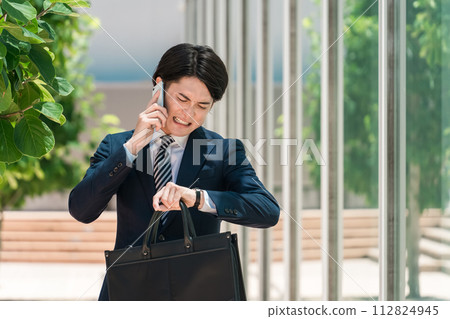 A young Asian male businessman in a suit is in a hurry, checking the time on his watch while talking on his smartphone. 112824945