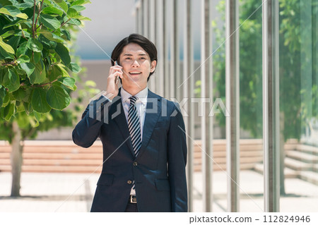 A young Asian male businessman in a suit talking on a smartphone while out on the road 112824946