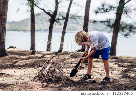 Cute boy digging soil in forest near sea beach. Family natural education skills. Travelling together. Learning camping organizing bonfire Cute boy digging soil in forest near sea beach. Family natural education skills. Travelling together. Learning camping organizing bonfire 112827460