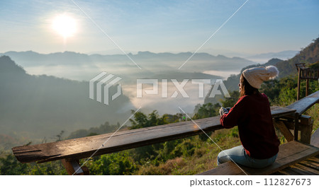 Asian female tourist is sitting and enjoying the morning sunrise and mist on the mountain. 112827673