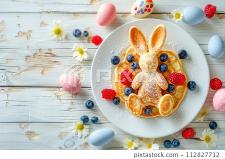 Easter holiday children's breakfast pancake in the shape of an Easter bunny with berries and Easter colored eggs on a light wooden background 112827712