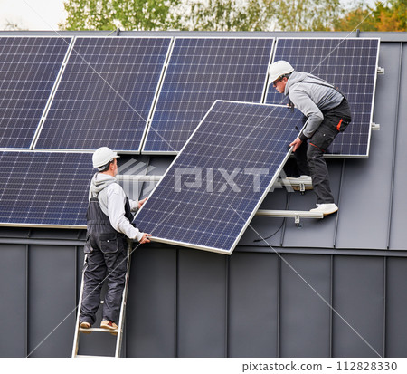 Mounters installing solar panel system on roof of house. Men workers in helmets carrying photovoltaic solar module outdoors. Concept of alternative and renewable energy. Mounters installing solar panel system on roof of house. Men workers in helmets carrying photovoltaic solar module outdoors. Concept of alternative and renewable energy. 112828330