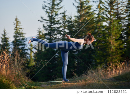 Middle-aged woman practicing yoga in forest. Sporty woman doing warrior 3 pose. Practicing gymtastic at dawn. 112828331