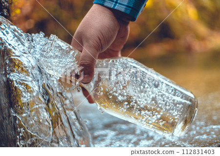 A hand brings a transparent bottle to a spring of drinking water. A man collects mineral water from a spring in the autumn forest. The traveler stopped at a spring to drink and collect clean water. A hand brings a transparent bottle to a spring of drinking water. A man collects mineral water from a spring in the autumn forest. The traveler stopped at a spring to drink and collect clean water. 112831403