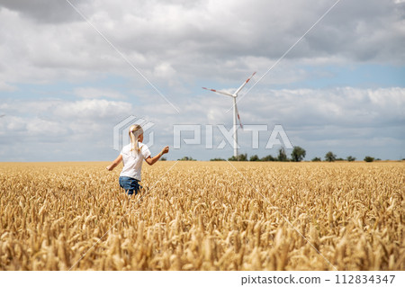 Back view little cute happy blond caucasian girl enjoy walking ripe wheat ear field against wind mill turbine farm on warm sunny summer day. Child freedom future clean energy environment concept 112834347
