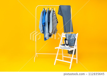 Denim pants and shirts on hangers, shoes on chair on yellow background Denim pants and shirts on hangers, shoes on chair on yellow background 112835645