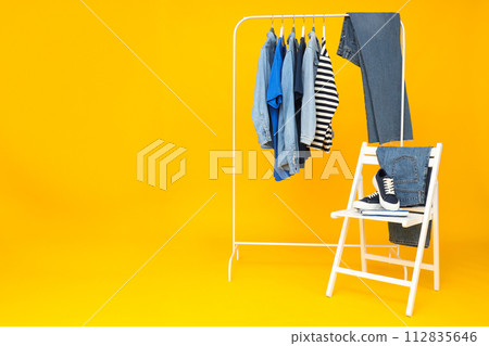 Denim pants and shirts on hangers, shoes on chair on yellow background, space for text Denim pants and shirts on hangers, shoes on chair on yellow background, space for text 112835646