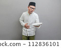 Asian Muslim man wearing a koko shirt and peci with shades of the fasting month, holding back hunger and plate while waiting for the time to break the fast, isolated on a gray background 112836101