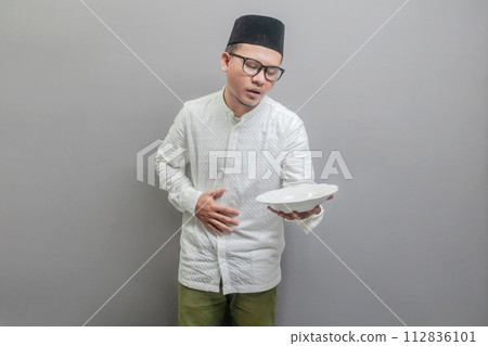 Asian Muslim man wearing a koko shirt and peci with shades of the fasting month, holding back hunger and plate while waiting for the time to break the fast, isolated on a gray background 112836101