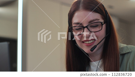 Closeup woman face wearing headset and talking, call center worker. Positive young woman wearing headset. Redhead woman holding meeting with colleagues in home interior. Closeup woman face wearing headset and talking, call center worker. Positive young woman wearing headset. Redhead woman holding meeting with colleagues in home interior. 112836720