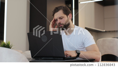 Tired man is working on laptop computer at the desk in kitchen at home office. Man upset stressed working at the laptop reading bad news. Concept of remote work or education. Tired man is working on laptop computer at the desk in kitchen at home office. Man upset stressed working at the laptop reading bad news. Concept of remote work or education. 112836843