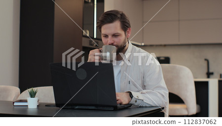 Young bearded man at home sitting in kitchen using laptop for distance work and drinking coffee. Online shopping, watching videos or writing emails. Young bearded man at home sitting in kitchen using laptop for distance work and drinking coffee. Online shopping, watching videos or writing emails. 112837062