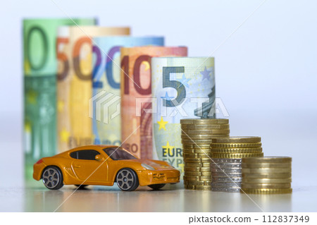 Blurred row of rolled hundred, fifty, twenty, ten and five new euro banknotes and pile of coins with yellow toy expensive sport car. Symbol of financial prosperity, vehicle sale and purchase. Blurred row of rolled hundred, fifty, twenty, ten and five new euro banknotes and pile of coins with yellow toy expensive sport car. Symbol of financial prosperity, vehicle sale and purchase. 112837349