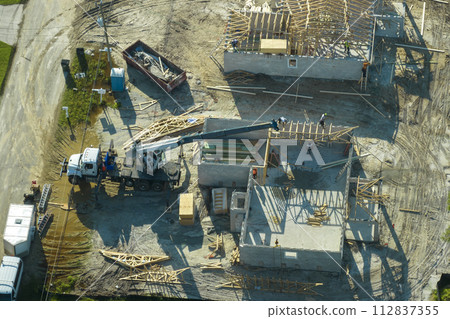 Aerial view of lifting crane and builders working on unfinished residential house with wooden roof frame structure under construction in Florida suburban area 112837355