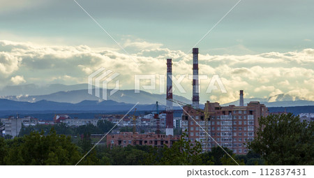 Wide panorama of modern urban building site of residential apartments quarter in city with green trees, working tower cranes, high pipe chimneys under blue sky on distant mountain range background. 112837431