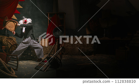 Clown in black hat, white face with red nose and striped pants playing over dark retro circus backstage background. 112838810